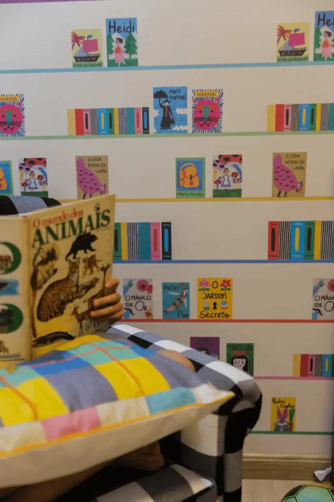 Menino lendo o livro O Mundo dos Animais em poltrona no cantinho da leitura com parede decorada de livros infantis.