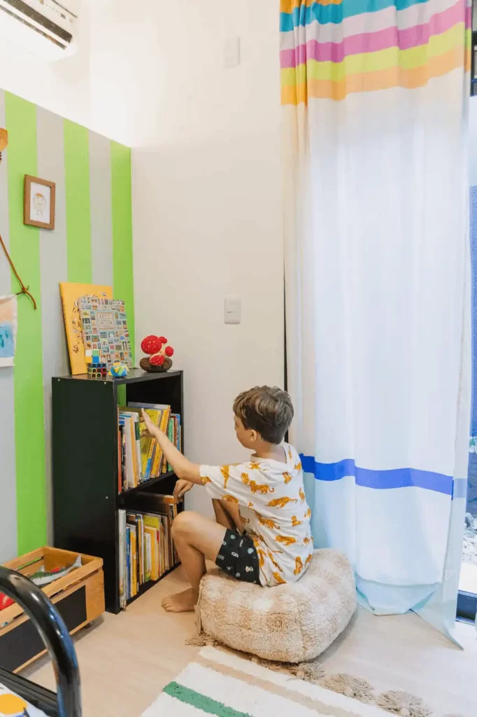 Menino sentado em almofada escolhendo livro infantil em uma estante no cantinho da leitura.