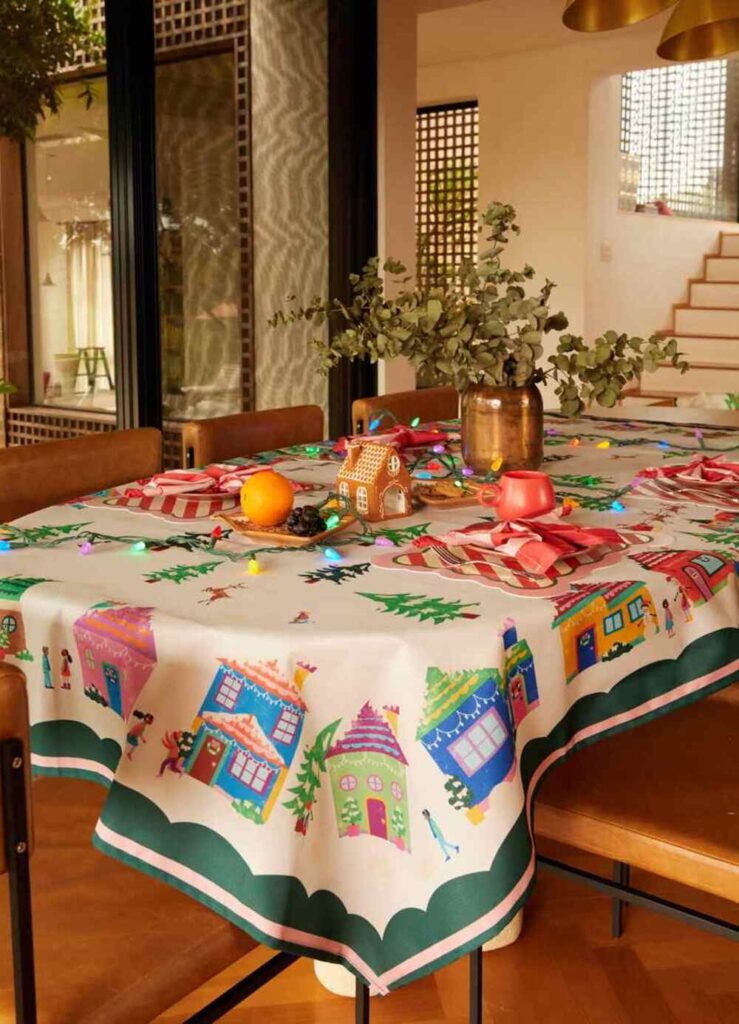 Mesa de jantar com toalha natalina colorida, casas ilustradas e enfeites festivos, decoração alegre para o Natal e Black Friday.