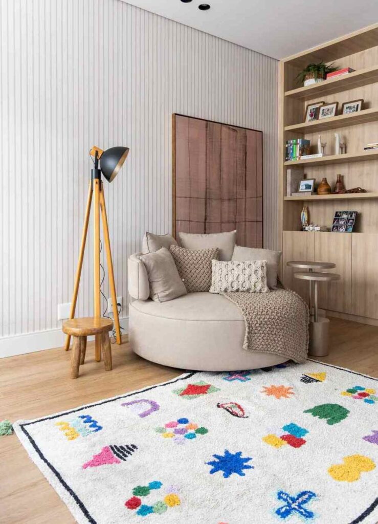 Quarto moderno com sofá redondo bege, almofadas de tricô e tapete colorido, decoração aconchegante com toque artesanal.