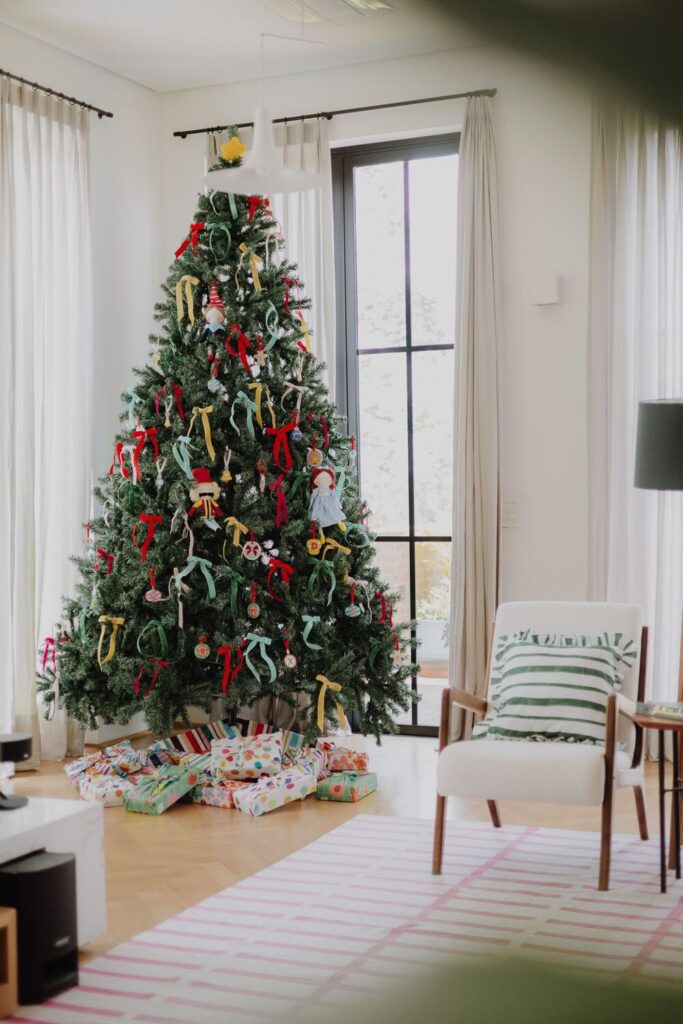 Árvore-de-natal com lacinhos e uma boneca em miniatura, com caixas de presente abaixo e cadeira ao lado