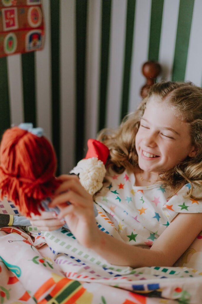 Menina sentada em cama sorrindo mexendo em boneca de pano