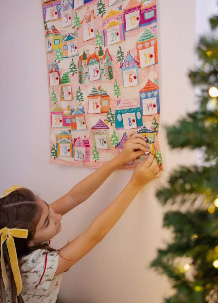 Menina colocando peças no calendário do Advento colorido com casas natalinas e árvore de Natal ao lado.