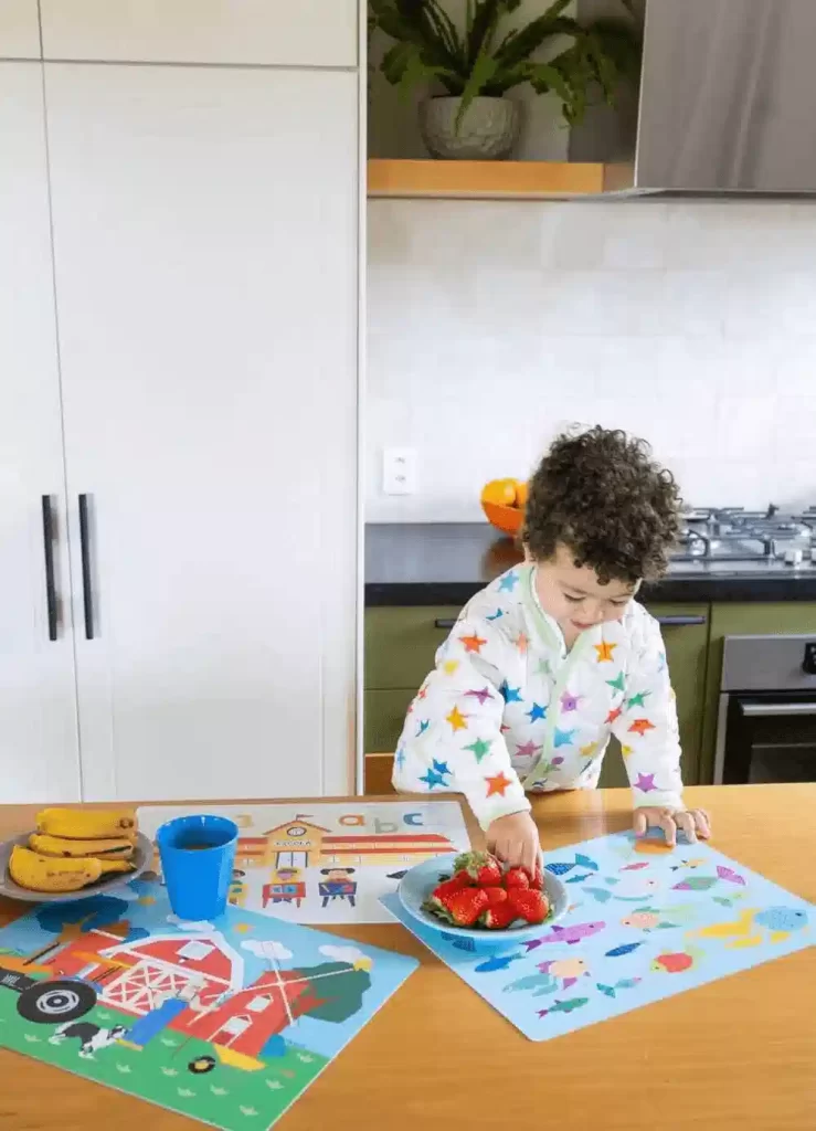 Criança ajudando na cozinha e pegando morangos em uma tigela azul sobre a mesa com frutas e jogos educativos.