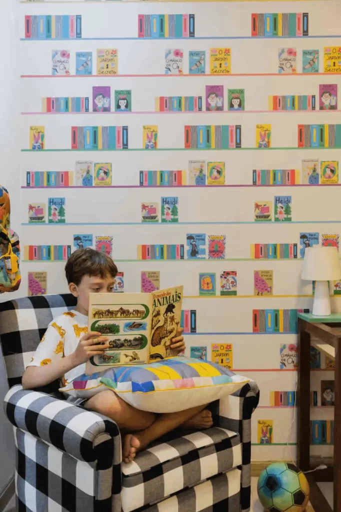 Menino sentado em poltrona xadrez lendo livro sobre animais, com parede decorada com capas de livros infantis