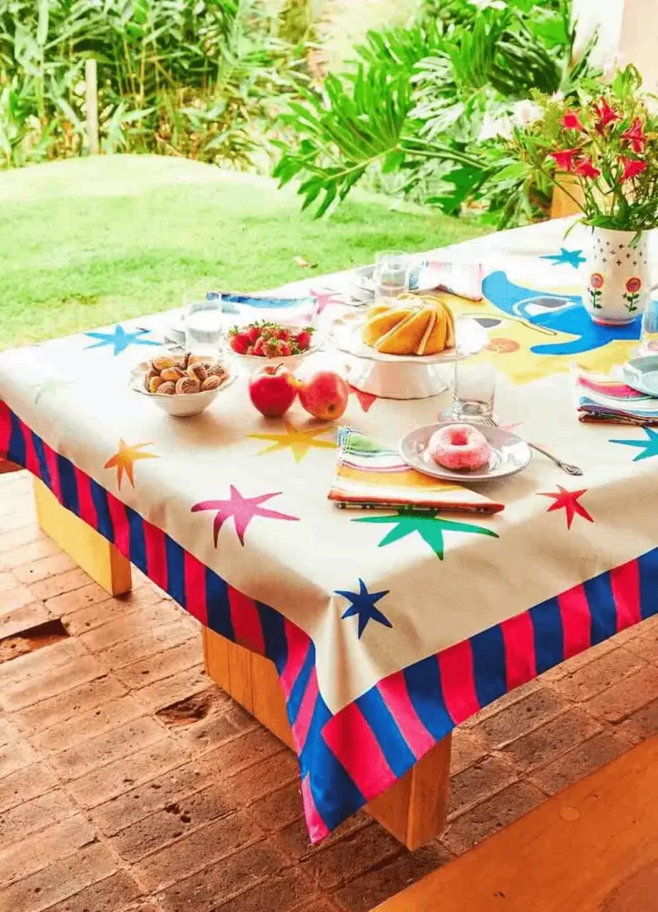 Crianças na cozinha ou área externa com mesa posta colorida, toalha estampada, frutas e bolo em ambiente familiar.