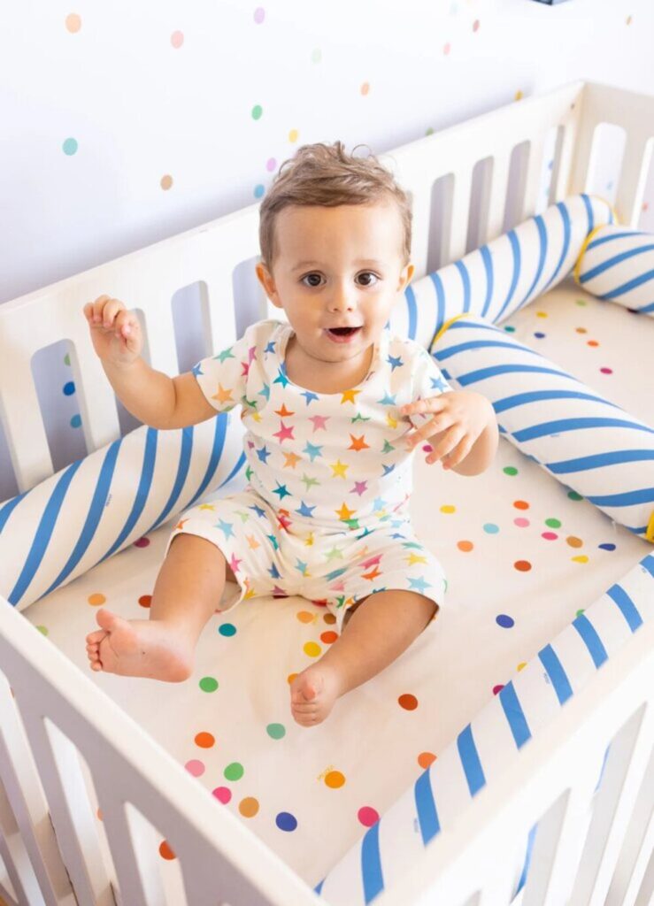 Bebê sentado em berço branco com protetores de berço listrados em azul e roupa estampada com estrelas coloridas.