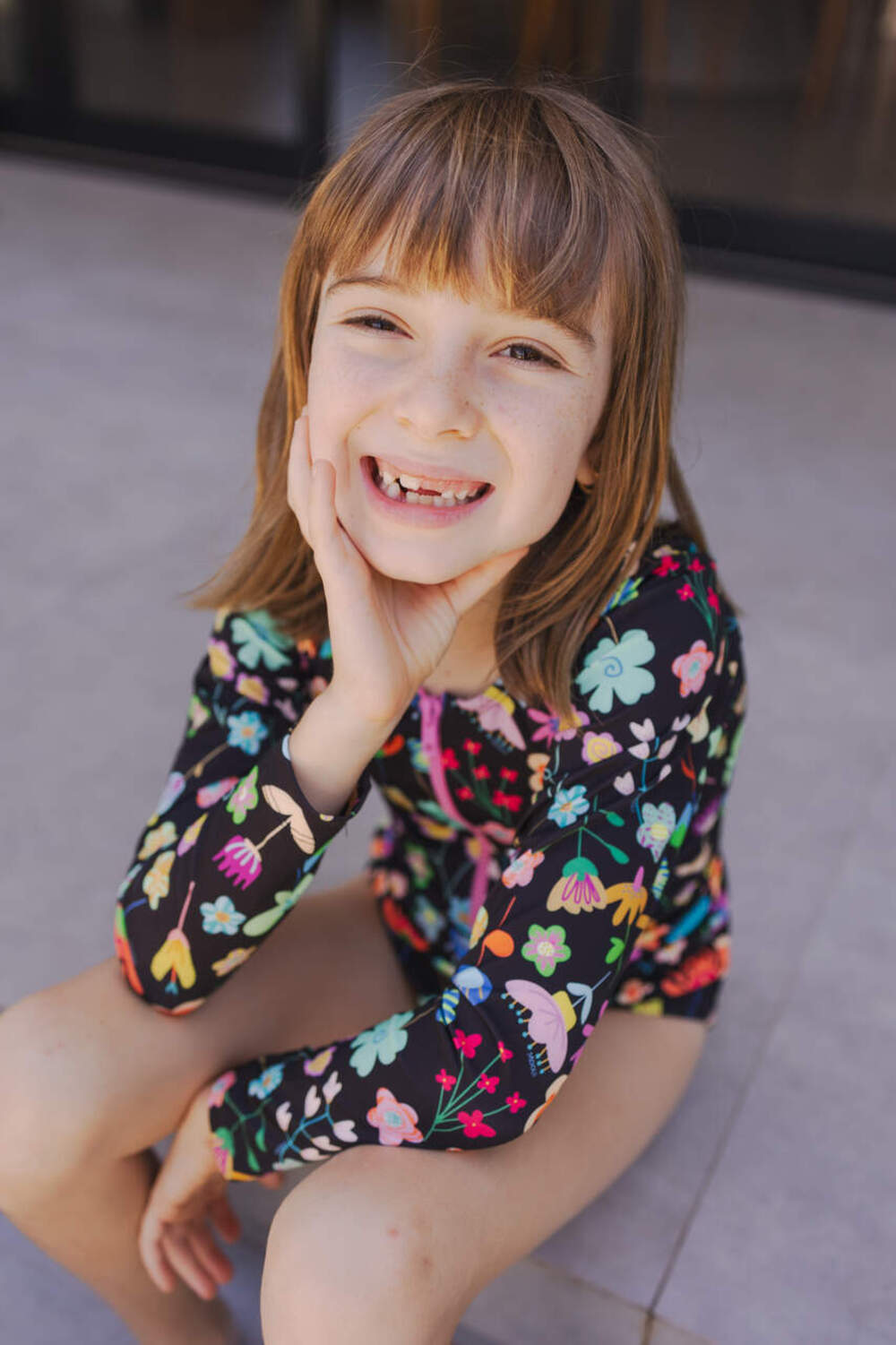 Menina sorridente com maiô floral de manga longa sentada no chão, em clima de verão.