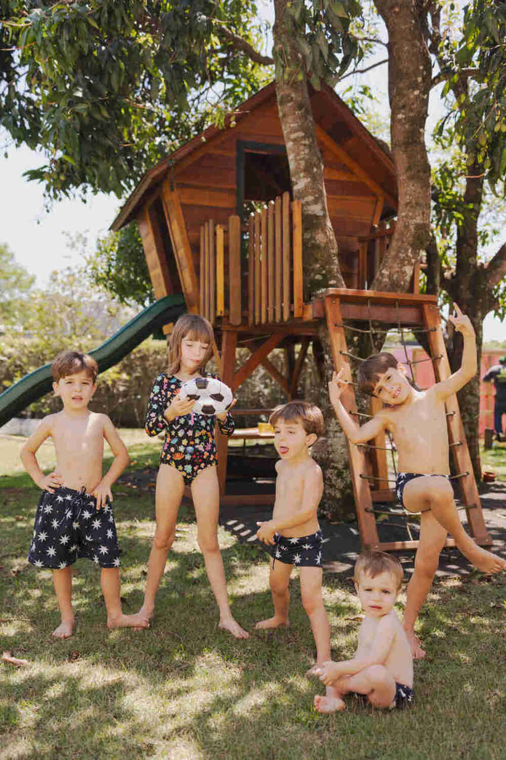 grupo de irmãos com roupas de banho escuras e estampadas posando felizes no quintal com casa na árvore