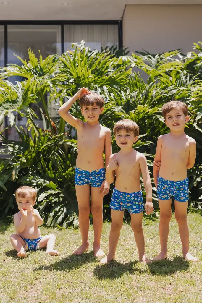 quatro meninos de sunga azul estampada se divertindo ao sol, em frente a plantas tropicais