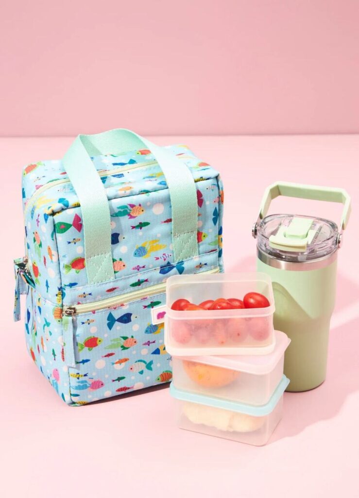 Lancheira térmica infantil de peixinhos para armazenar hambúrguer de atum com potes de frutas e copo térmico sobre fundo rosa.