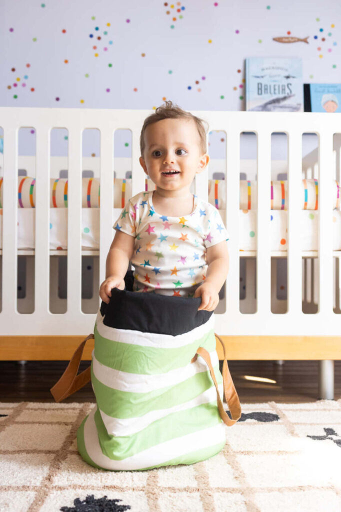 Bebê sorrindo em pé dentro de um saco de dormir listrado verde e branco no quarto infantil.