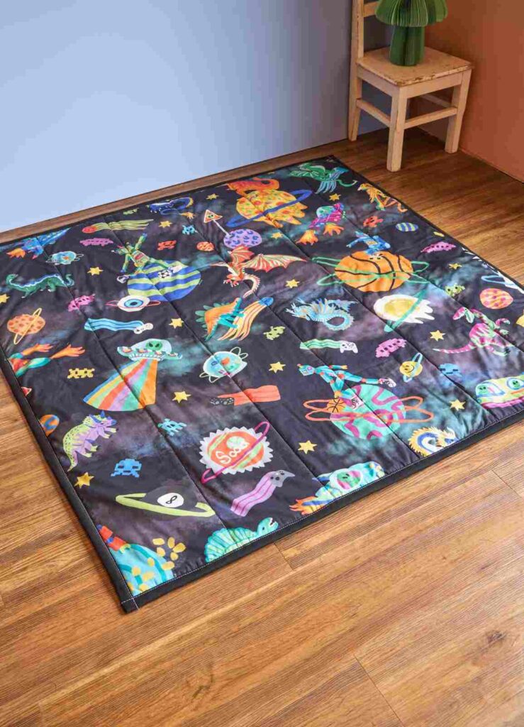 Tapetinho de brincar acolchoado com estampa espacial colorida, ideal para bebês explorarem com conforto e segurança.